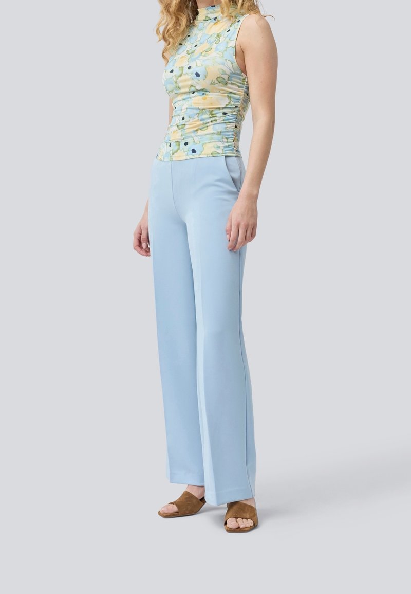 Femme portant un haut fleuri sans manches, un pantalon large bleu clair et des sandales ouvertes marron, debout devant un fond uni.