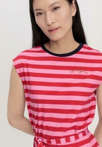 Gestreifter roter und rosa ärmelloser Top mit einem dunklen Halsauschnitt, der "imagine" in Gold gestickt hat. Mit einem Knoten an der Taille für eine taillierte Optik.