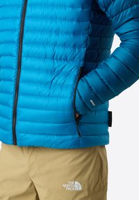 Blå isolerad jacka med kviltad mönster, svart dragkedja och sidofickor. Kombinerad med ljusbeige shorts med en vit logotyp.