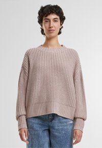 Rosa gestrickter Pullover mit rundem Halsausschnitt, gerippten Bündchen und lockerer Passform. Gewebte Textur und überschnittene Schultern ergänzen die legere Jeans.