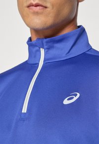 Blå halv-zip genser laget av glatt stoff, med en hvit glidelås og et logo på brystet. Krage-design med sydde detaljer.