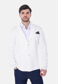 Cardigan bianco a maglia con due tasche frontali e bottoni in legno chiaro. Indossato sopra una camicia con colletto, con un fazzoletto da taschino decorato.