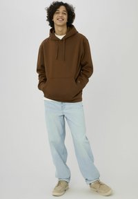 Jeune homme debout, souriant, portant un sweat à capuche marron, un jean bleu clair et des chaussures beige sur un fond uni gris clair.