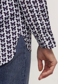 Skjorta med knappar framtill i lätt tyg med vit bakgrund och lila fjärilstryck, rundad nederkant och enknappsknappar vid ärmarna.