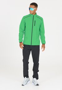 Veste coupe-vent verte avec des accents noirs, poche zippée, associée à un pantalon noir et des baskets grises. Des lunettes de soleil réfléchissantes complètent le look.