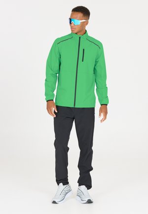 Grön vindjacka med svarta detaljer, ficka med dragkedja, kombinerad med svarta byxor och grå sneakers. Reflexglasögon fulländar looken.