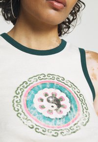 Person wearing white tank top with dark green trim and a faded yin-yang symbol surrounded by floral and swirl patterns on the chest.