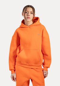 Sudadera con capucha naranja de tejido suave, con un bolsillo frontal, puños de canalé y un pequeño detalle de logo en el pecho. Combinada con pantalones a juego.
