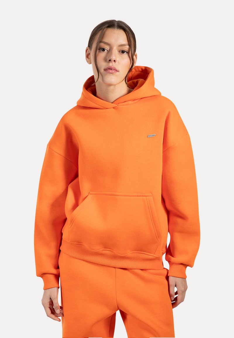Sudadera con capucha naranja de tejido suave, con un bolsillo frontal, puños de canalé y un pequeño detalle de logo en el pecho. Combinada con pantalones a juego.