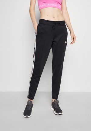 Vrouw draagt zwarte Adidas trainingsbroek met witte strepen, gecombineerd met zwarte Adidas sneakers en een roze crop top.