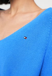 Modrý pletený sveter s textúrovaným vzorom, na ľavom ramene s malým logom vlajky v červenej, bielej a modrej farbe.