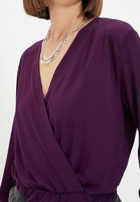 Purple wrap blouse with long sleeves, made from smooth fabric. Features a deep V-neck and a gathered waistband, complemented by a layered silver necklace.
