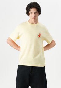Camiseta de algodón amarilla con mangas cortas, escote redondo y un pequeño diseño gráfico rojo en el pecho, combinada con pantalones de mezclilla oscuros.