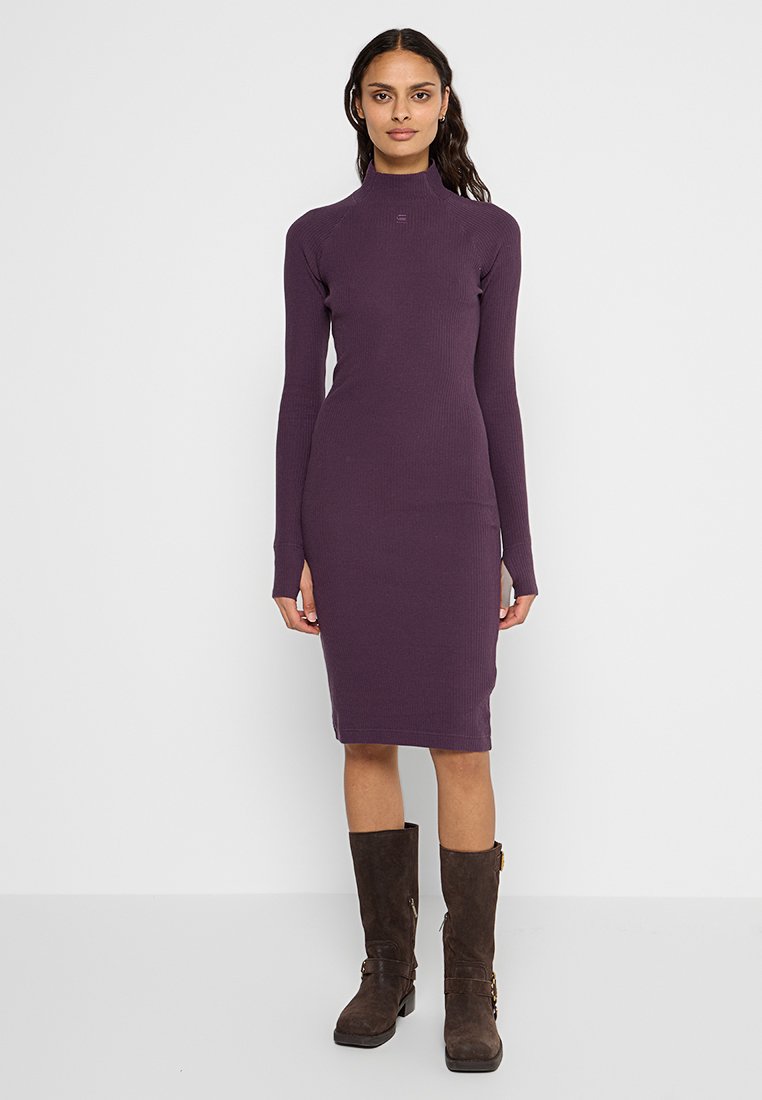 Fioletowa, żeberkowana sukienka z wysokim dekoltem i długimi rękawami, dopasowana sylwetka, sięgająca do połowy łydki. Modelka ma na sobie wysokie brązowe kozaki.