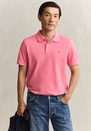 Mann trägt ein pinkes Poloshirt und blaue Jeans, hält eine dunkle Jacke in einer Hand und steht vor einem schlichten hellen Hintergrund.