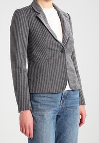 Veste à rayures grises avec col cranté, fermeture à un bouton et deux poches avant, portée sur une chemise blanche et un jean bleu.