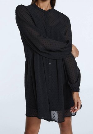 Vrouw die een zwarte doorschijnende jurk met stippen draagt, lange mouwen met elastische manchetten, staande tegen een lichte achtergrond.