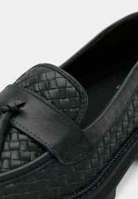 Loafers din piele neagră, cu o textură împletită, având un vârf rotund și neted, precum și o curea decorativă cu detaliu înnodat în partea din față.