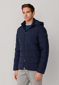 Chaqueta acolchada azul marino con capucha, con costuras horizontales, bolsillos laterales con cremallera y cremallera frontal completa. Llevada sobre un suéter azul.