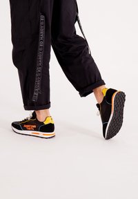 Sneaker nere e gialle con una suola bianca e arancione, dotate di una tomaia in rete e testo "DOWNTOWN TOKYO" in bold arancione. Design a taglio basso.