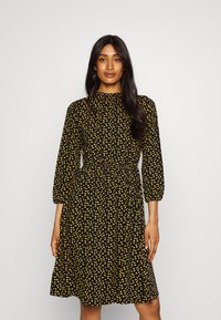 Veste preta com bolinhas amarelas, mangas longas, decote redondo e uma cintura elástica que se alarga até o comprimento do joelho.