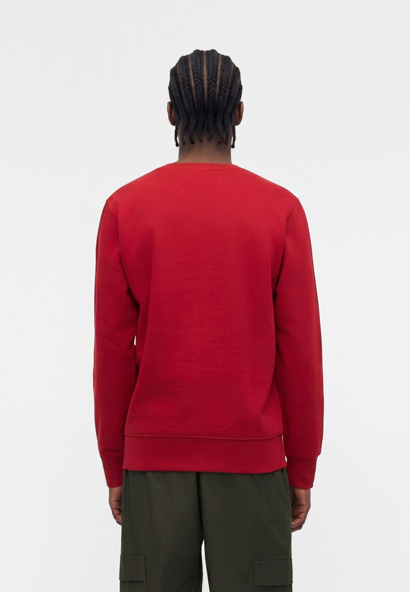Persona con el pelo trenzado que lleva una camisa roja de manga larga y pantalones cargo verde oscuro, de espaldas sobre un fondo claro y liso.