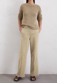 Femme debout contre un mur blanc, portant un pull en tricot beige, un pantalon ample beige et des sandales noires avec des boucles dorées.