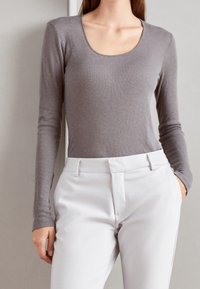 Kvinna klädd i en åtsittande grå långärmad topp, instoppad i högmidjade vita byxor med en hand i fickan.