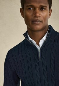 Marinblå stickad tröja med kabelmönster, hög krage och halvdragkedja. Buren över en vit skjorta med krage, vilket framhäver textur och tonkontrast.