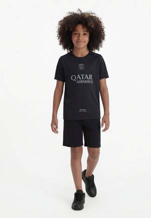 Enfant aux cheveux bouclés portant une tenue de sport noire du Paris Saint-Germain marchant vers l'avant sur fond blanc.