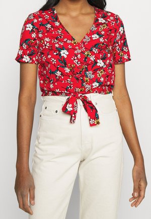 Femme portant un chemisier rouge à fleurs à manches courtes, noué à la taille, associé à un pantalon blanc taille haute sur un fond uni.