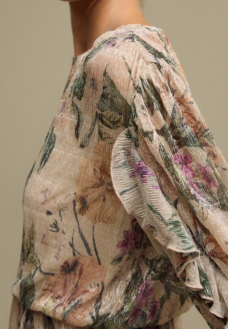 Gros plan d’un chemisier transparent à fleurs avec manches longues à volants dans des tons doux de rose, vert et violet sur une personne.