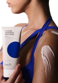 Siero corpo antiossidante in un tubo bianco con design circolare blu. Crema applicata sulla pelle; il soggetto indossa una maglietta senza maniche blu.