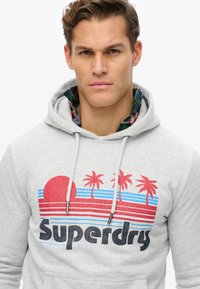 Jasnoszara bluza z kapturem i sznurkami, ozdobiona graficznym wzorem przedstawiającym czerwoną słońce, palmy, niebieskie i czerwone paski oraz tekst "Superdry".