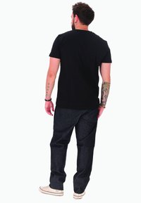 Uomo con tatuaggi su entrambe le braccia di spalle, indossa una t-shirt nera, jeans scuri, sneakers bianche e braccialetti sul polso sinistro.