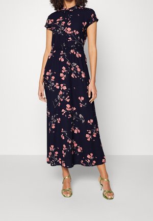 Kobieta ubrana w granatową sukienkę midi z krótkim rękawem, ozdobioną różowymi kwiatowymi wzorami, w połączeniu z metalicznie zielonymi sandałami na wysokim obcasie.