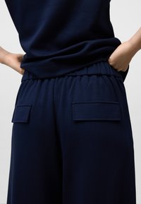 Persoon in marineblauwe elastische taillebandbroeken met achterzakken en een bijpassend marineblauw bovenstuk, handen op de heupen.