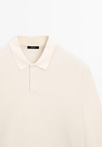 Cremfarbener Langarm-Polohemd mit zwei Knöpfen und umgeschlagenem Kragen, flach auf einem weißen Hintergrund ausgelegt.