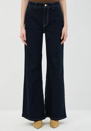 Dunkelblaue weitgeschnittene Jeanshose mit Kontrastnähten, hoher Taille und vorderen Taschen. Kombiniert mit beigen spitzen Pumps.