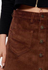 Jupe en velours côtelé marron avec nervures verticales, fermeture à boutons métalliques à l'avant, et une seule poche plaquée sur le côté.