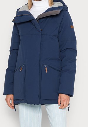 Frau trägt eine dunkelblaue wattierte Winterjacke mit Kapuze und Reißverschlusstaschen vorne, kombiniert mit hellen gestreiften Hosen.
