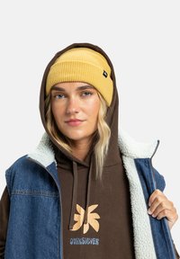 Ung kvinne som har på seg en gul strikket lue, en brun hettegenser med logo, og en blå denimvest med sherpa-fôr, ser inn i kameraet.