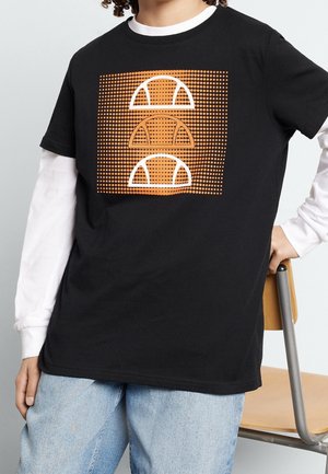 Personne portant un t-shirt noir avec un carré pointillé orange et trois formes semi-circulaires blanches et orange, superposé à une chemise blanche à manches longues.