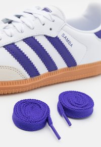 Vit sneaker med lila läderremsor och gummsula, med rund tå, texturerat material och två rullade lila skosnören.