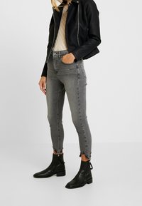 Szare, wysokie skinny jeansy z postrzępionymi nogawkami, połączone z czarną skórzaną kurtką i czarnymi botkami, wyeksponowane na neutralnym tle.