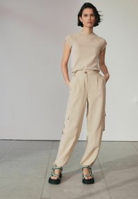 Beige kortærmet top med rund hals, parret med løstsiddende beige cargo bukser med sidelommer. Sorte sandaler med grønne detaljer.