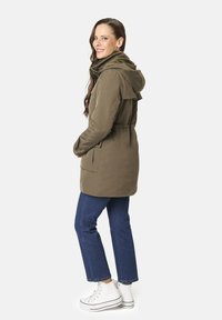 Parka verde oliva con capucha, que presenta una cintura ajustada, bolsillos delanteros y un corte relajado. Combinada con vaqueros azules y zapatillas blancas.