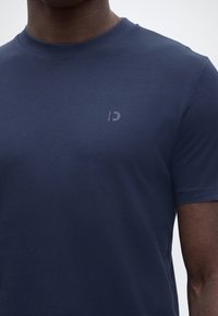 Κοντινό πλάνο ενός ατόμου που φοράει ένα μονόχρωμο μπλε σκούρο κοντομάνικο t-shirt με στρογγυλή λαιμόκοψη και ένα μικρό, διακριτικό λογότυπο στο αριστερό μέρος του στήθους.