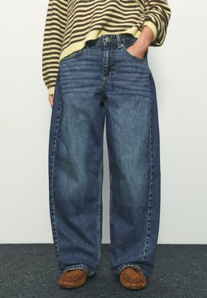 Oseba, ki nosi široke dvobarvne modre kavbojke z rokami v žepih, v kombinaciji z rjavimi čevlji v stilu mokasink in črtastim puloverjem.