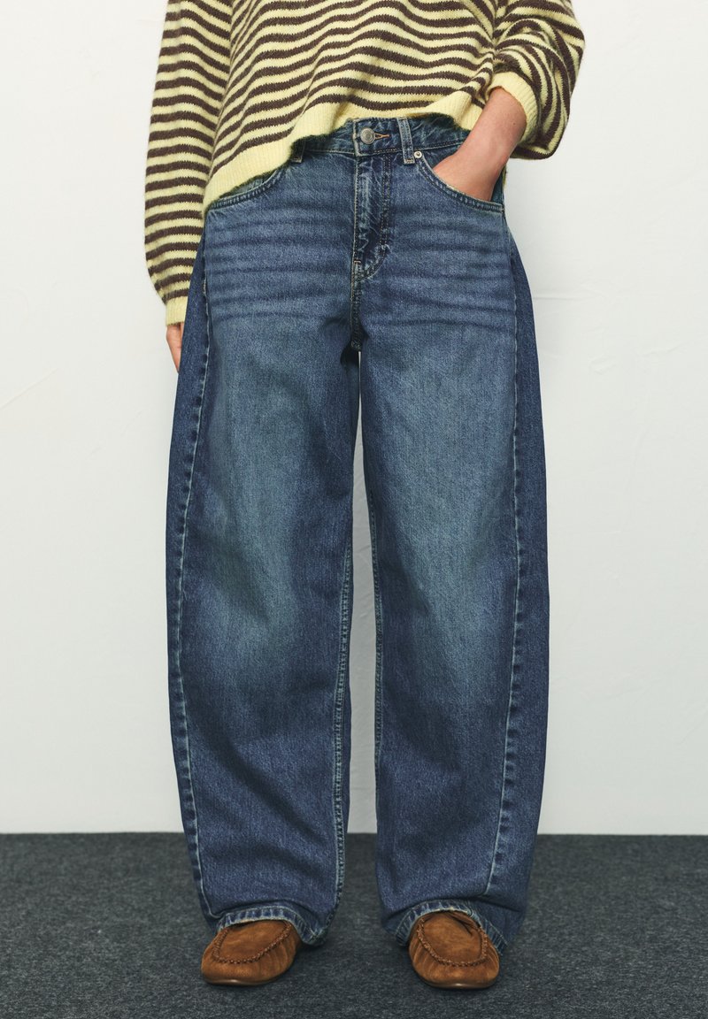 Personne portant un jean deux tons bleu à coupe ample, les mains dans les poches, associé à des mocassins marron et un pull rayé.